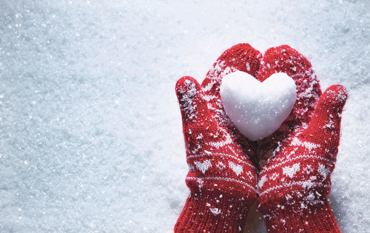 Über einer Schneedecke halten zwei rote Handschuhe ein Herz aus Schnee.