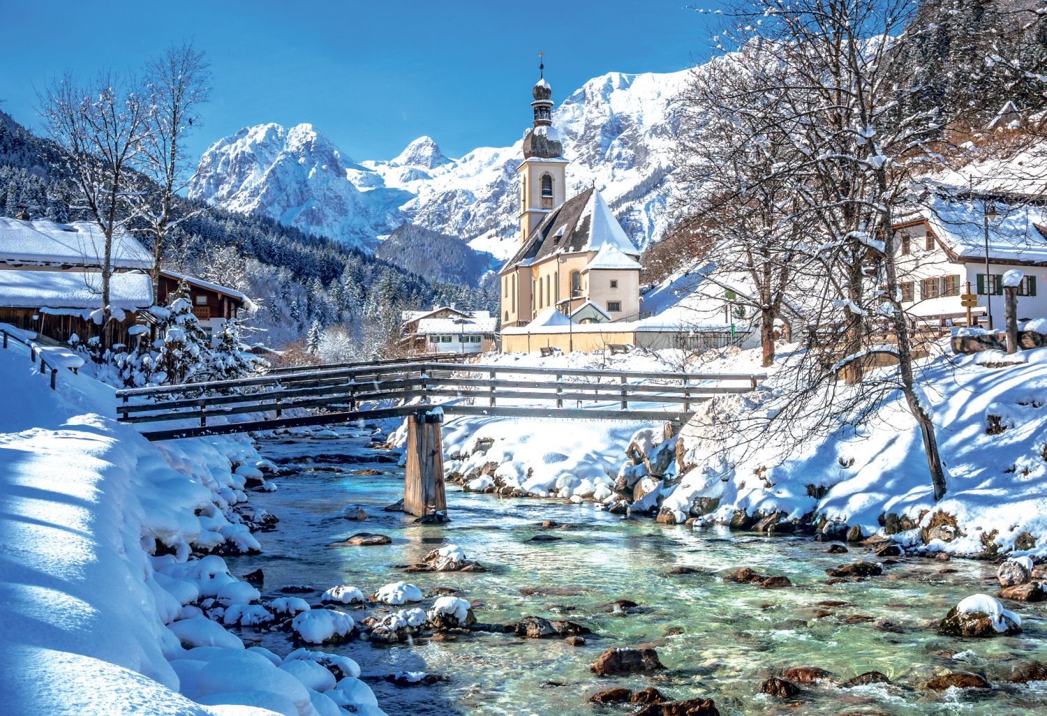 Eine Winterlandschaft mit einem eingeschneiten Dorf, einer Brücke und einem Fluss vor einem blauen Himmel.