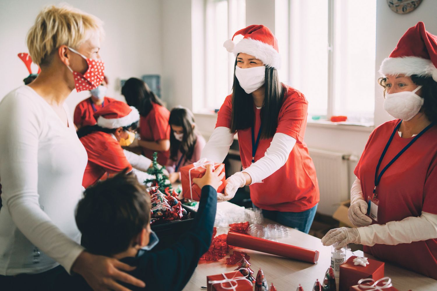 Eine Frau mit Weihnachtsmütze verteilt an eine Frau und ein Kind Geschenke. 